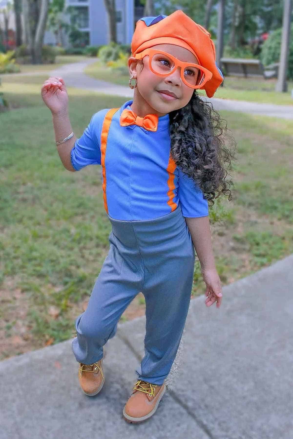 Child wearing blue shirt, orange suspenders, orange bow tie, orange hat, orange glasses, gray pants, and brown shoes, posing outdoors on a paved path with trees and grass in the background.