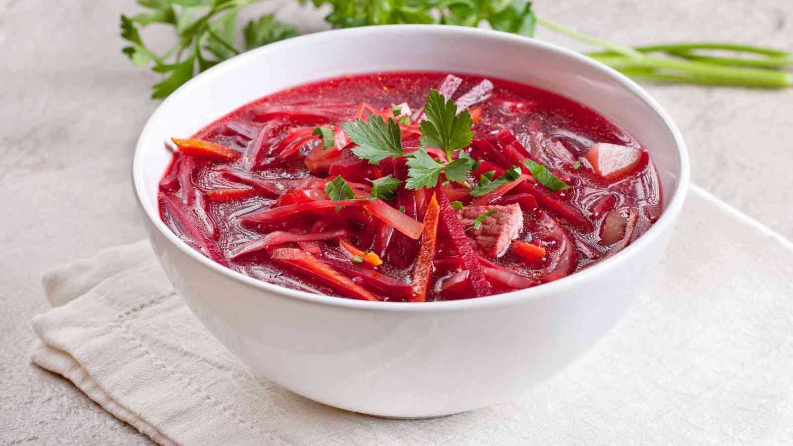 A white bowl filled with beet soup garnished with fresh parsley, placed on a light-colored cloth napkin with herbs in the background.