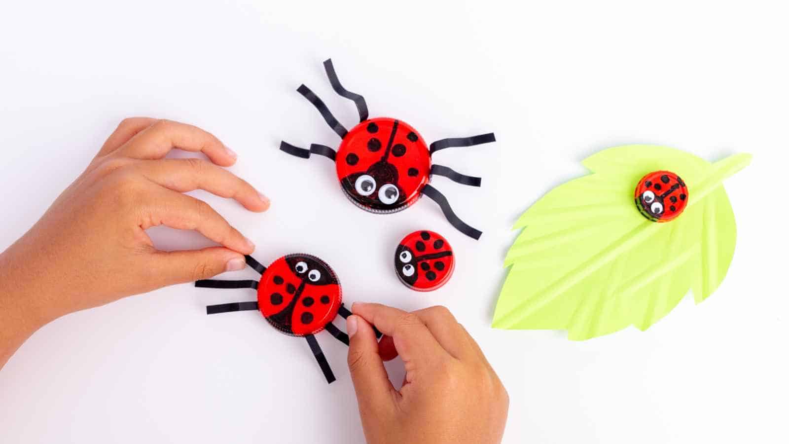 Two hands arrange red ladybug crafts made from bottle caps, googly eyes, and black paper legs, with one ladybug placed on a light green leaf-shaped surface.