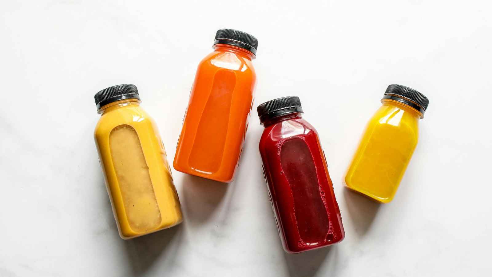 Four plastic bottles filled with orange, red, yellow, and beige juices or smoothies are arranged diagonally on a white surface.