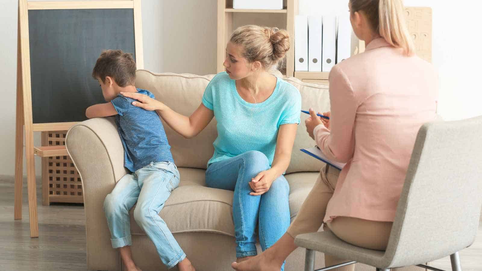 A woman comforts a young boy who is sitting on a couch with his face turned away, while another woman with a clipboard sits nearby, possibly in a counseling session.