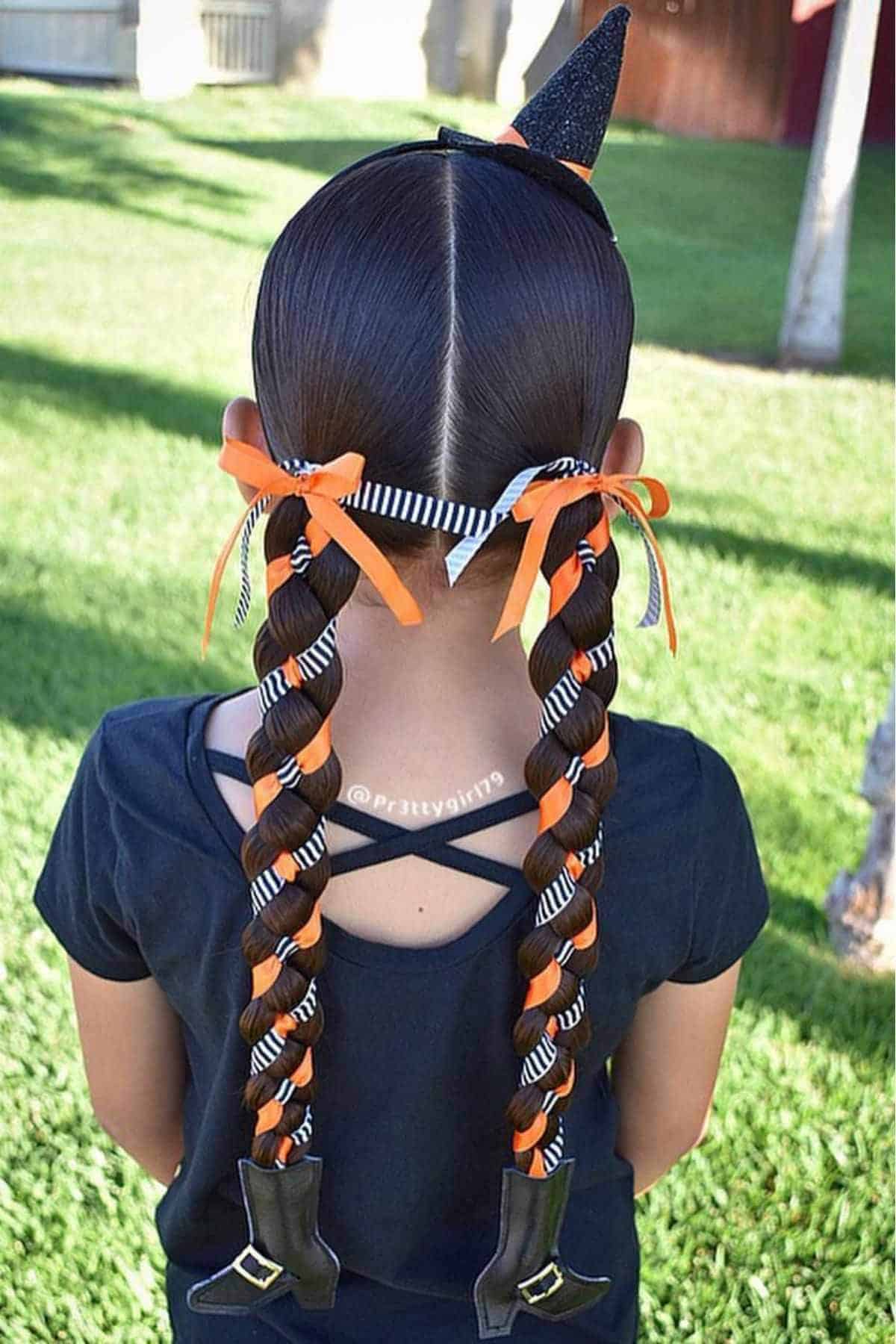 A girl with long braided hair decorated with orange and black ribbons, small witch hats at the ends, and a witch hat headband, standing outdoors on grass.
