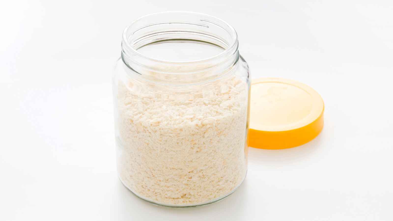 A clear glass jar filled with white powdered substance, possibly flour or grated cheese, sits on a white surface next to its yellow lid.