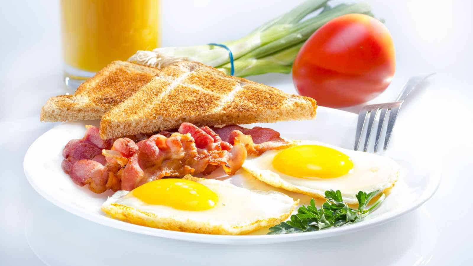 A plate with two sunny-side-up eggs, strips of bacon, and two slices of toast. In the background are a tomato, green onions, and a glass of orange juice.