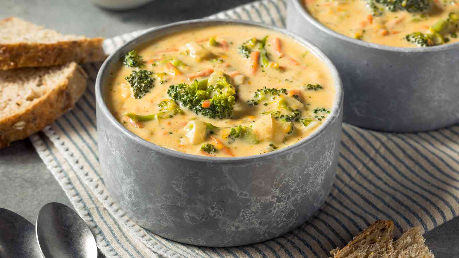 A bowl of creamy broccoli cheddar soup with visible broccoli florets and shredded carrots, served with slices of bread on the side.