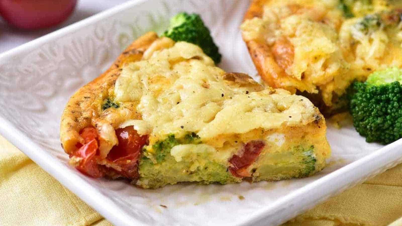 A slice of vegetable frittata with visible broccoli and tomatoes sits on a white plate.