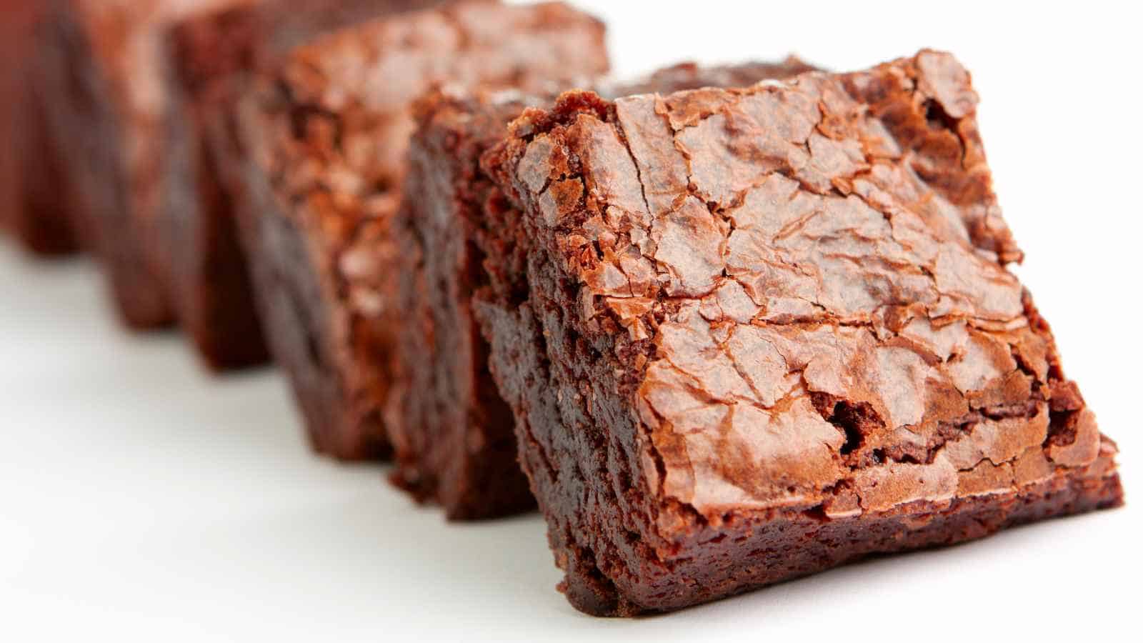 Close-up of several chocolate brownies arranged in a row on a white surface, showing a flaky, cracked top and dense, rich interior.