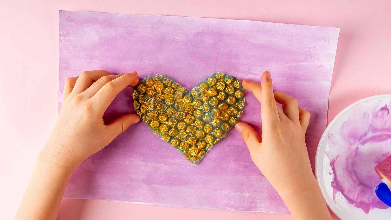 Two hands hold a heart-shaped piece of painted bubble wrap on a purple watercolor background, with a plate containing paint nearby.