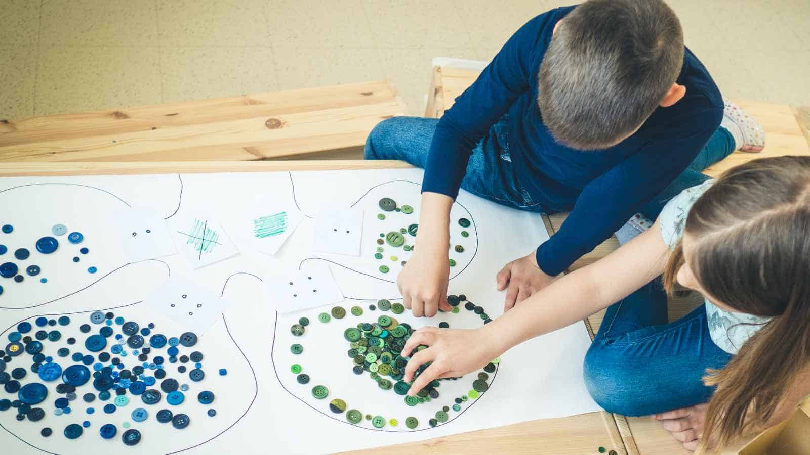 Two children sort blue and green buttons into sections drawn on a large sheet of paper, sitting on a wooden floor.