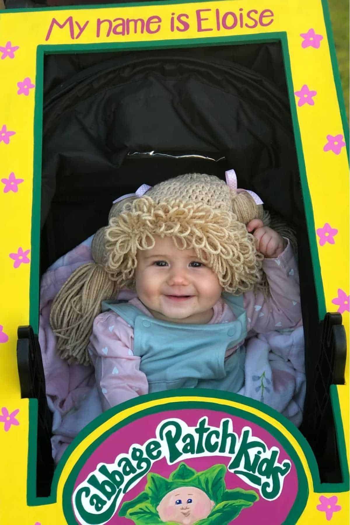 A baby dressed as a Cabbage Patch Kid sits in a decorated box labeled "My name is Eloise" and "Cabbage Patch Kids," wearing a yarn wig and smiling.