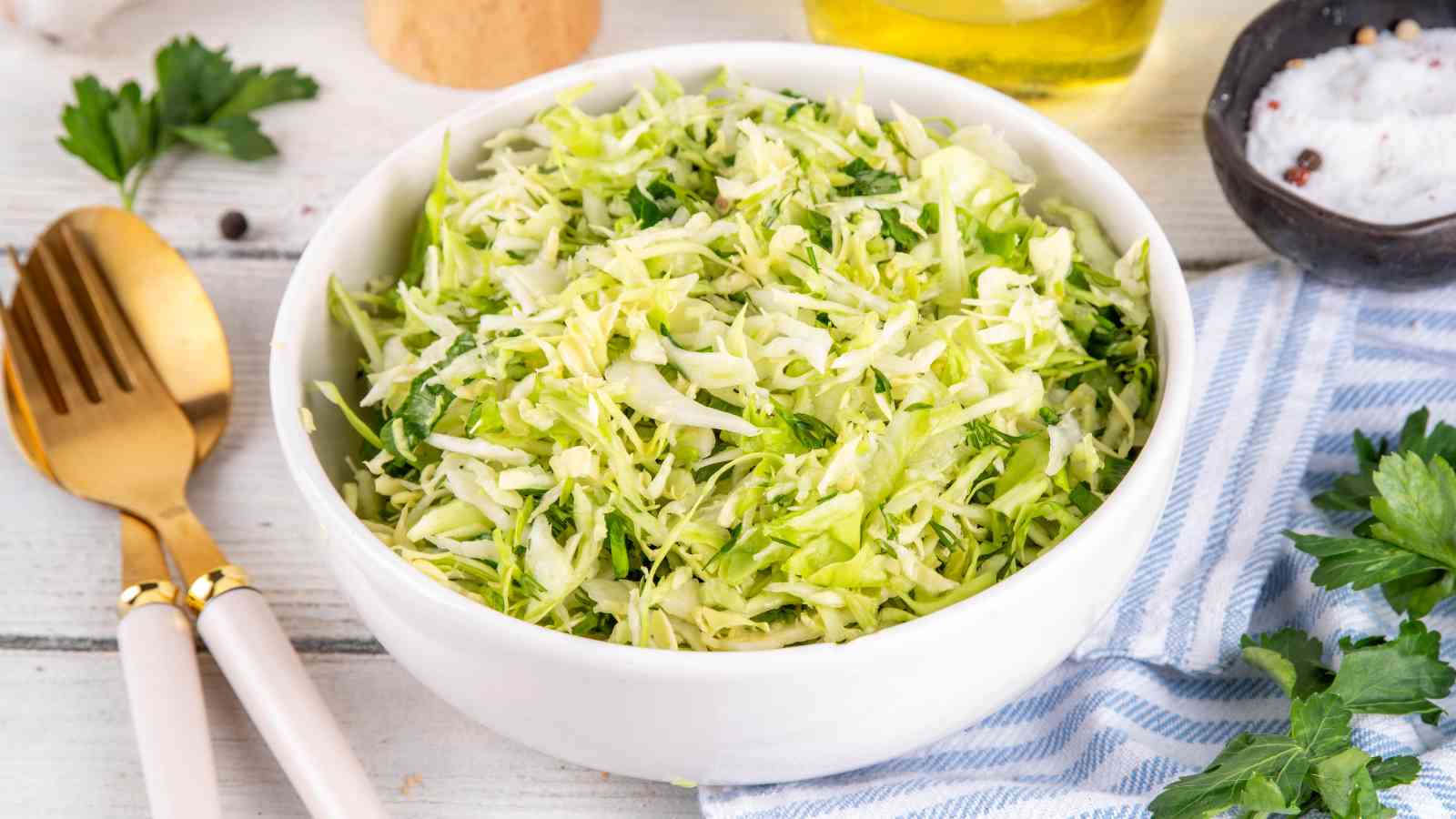 A white bowl filled with shredded cabbage salad sits on a table next to gold utensils, fresh parsley, and a striped napkin.