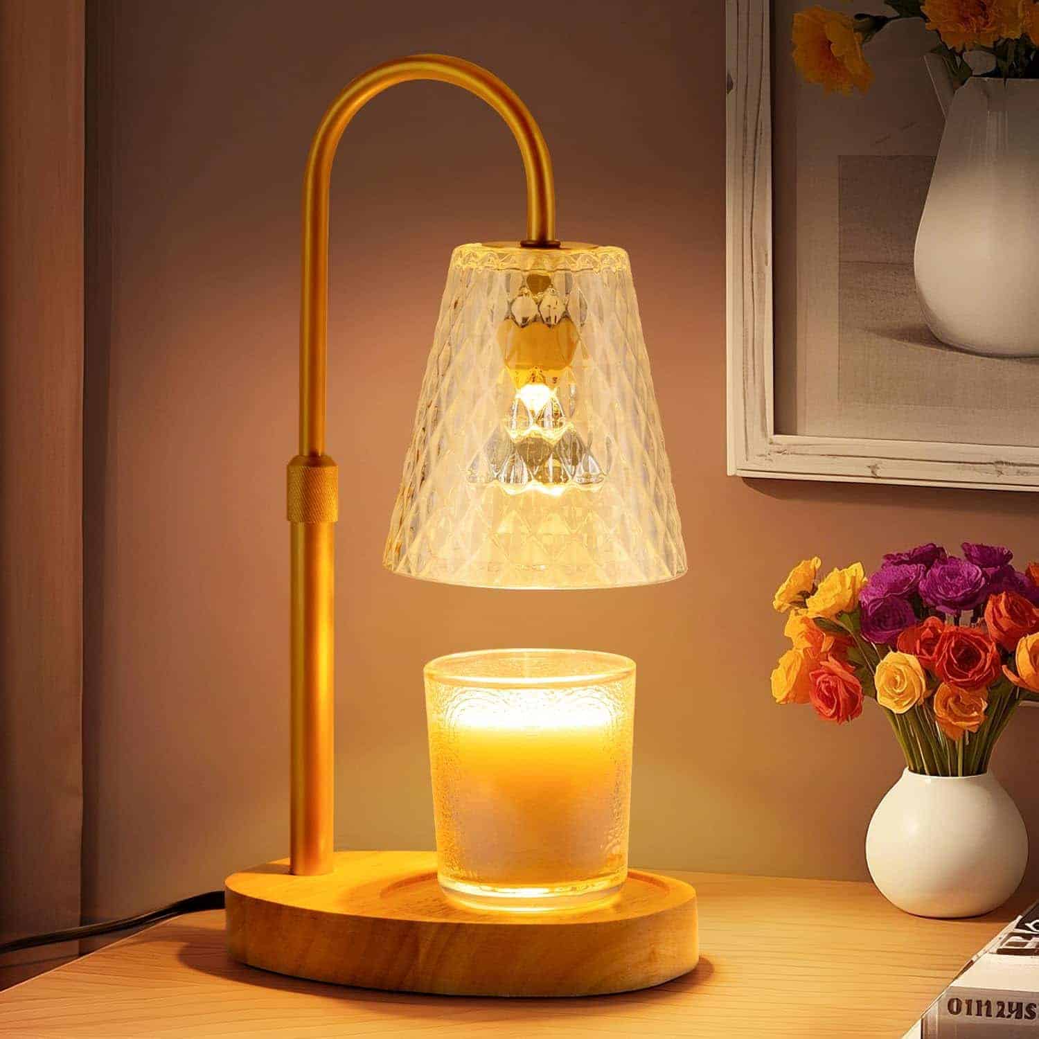 A gold lamp with a textured glass shade illuminates a candle on a wooden base, placed on a desk beside a vase of flowers and a framed picture.