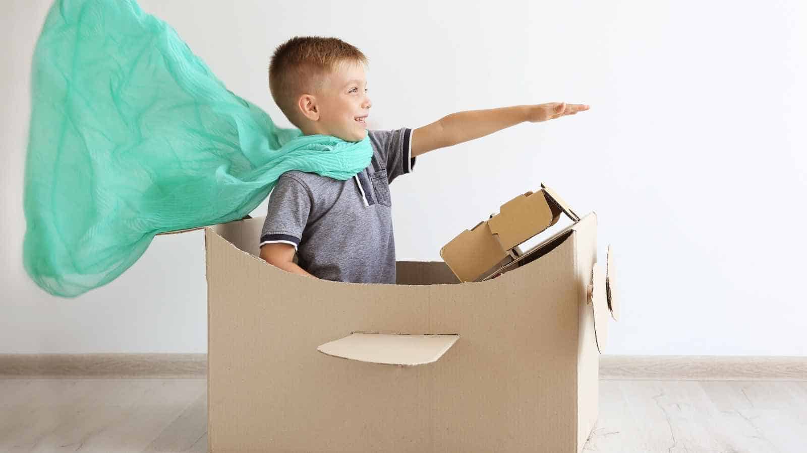 A young boy sits in a cardboard box shaped like a car, wearing a light blue scarf as a cape and extending his arm forward, pretending to drive or fly.