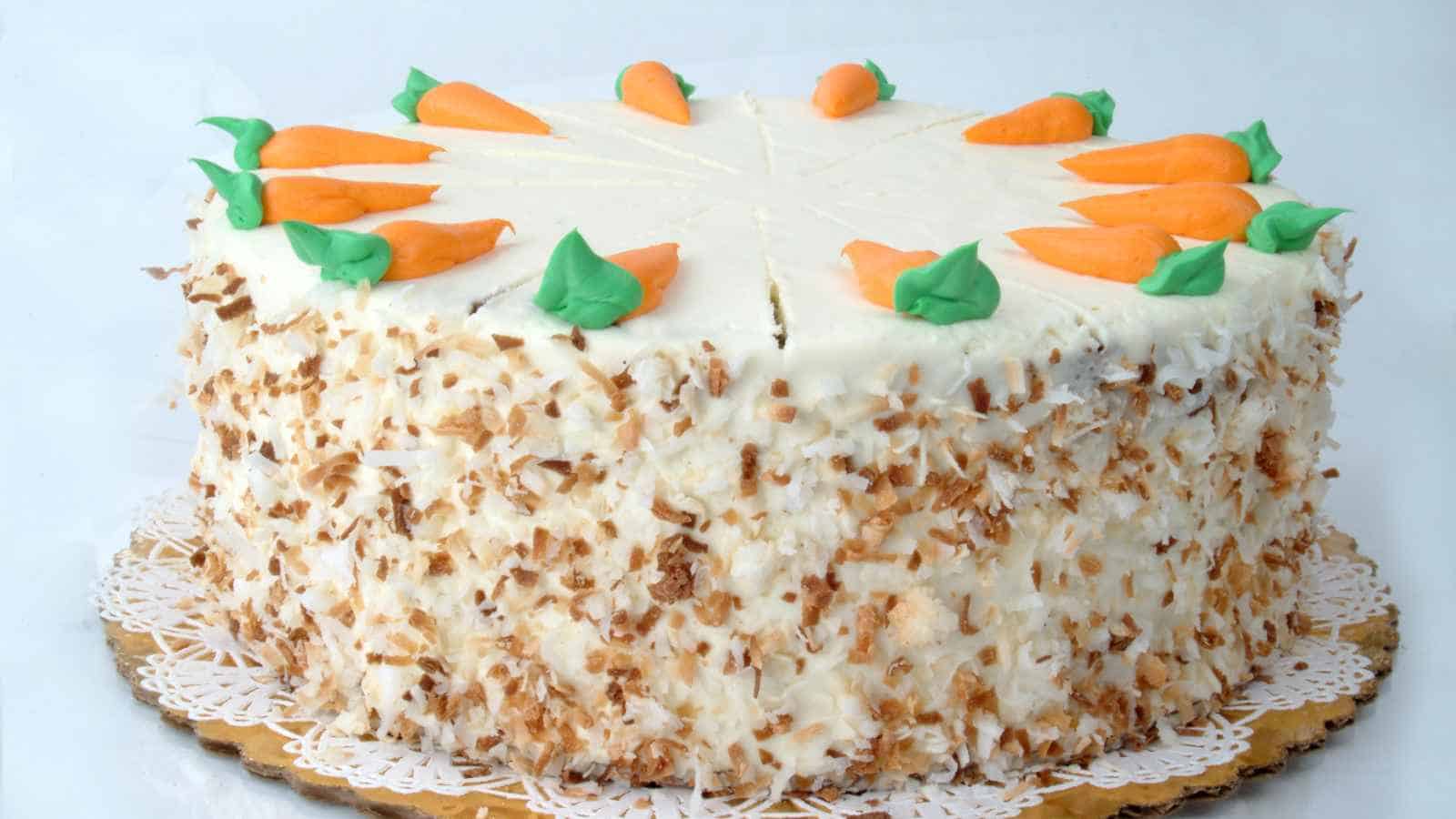 Round carrot cake with white frosting, shredded nuts on the sides, and decorative orange and green icing carrots on top, placed on a lace paper doily.