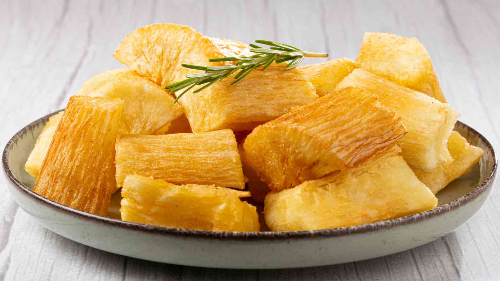 A plate of fried yuca (cassava) pieces garnished with a sprig of rosemary, served on a light-colored dish.