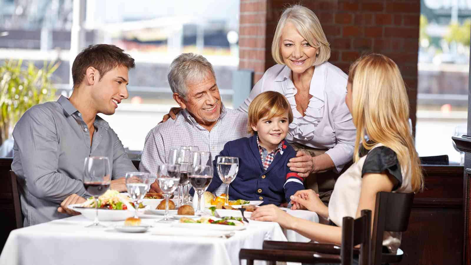 A family of five, including a young boy, gathers around a restaurant table with food and drinks, smiling and engaging in conversation.