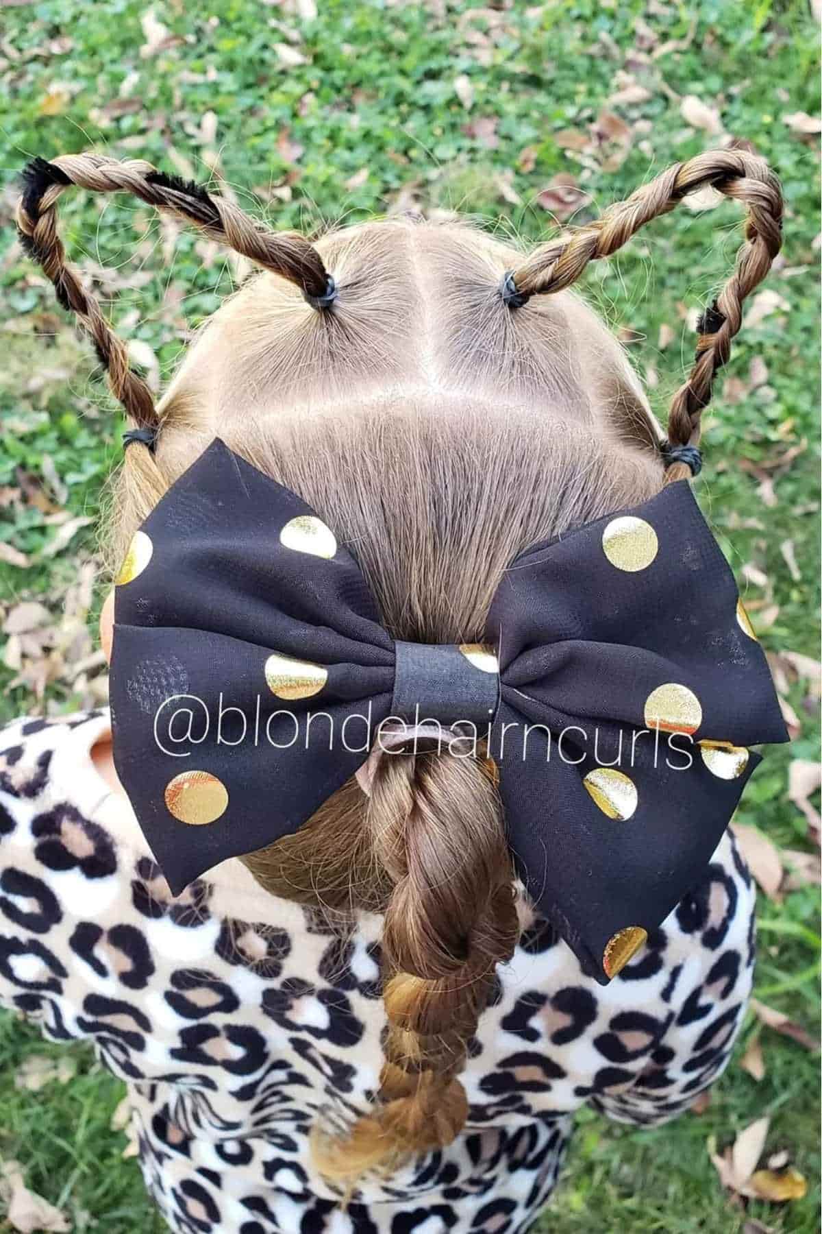 A child with braided hair styled into cat ears, wearing a large black bow with gold polka dots and a leopard print top, seen from behind.