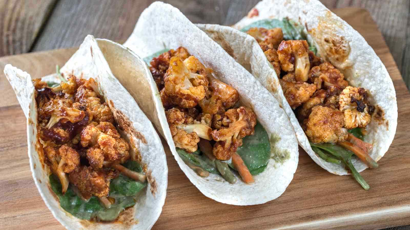 Three soft tacos filled with roasted cauliflower, leafy greens, and shredded vegetables on a wooden board.