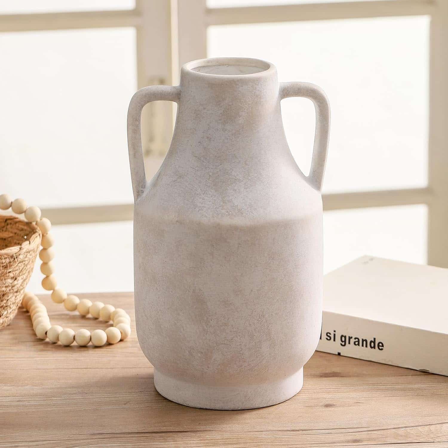 A white ceramic vase with two handles sits on a wooden table next to a beaded necklace, a woven basket, and a book labeled "si grande.