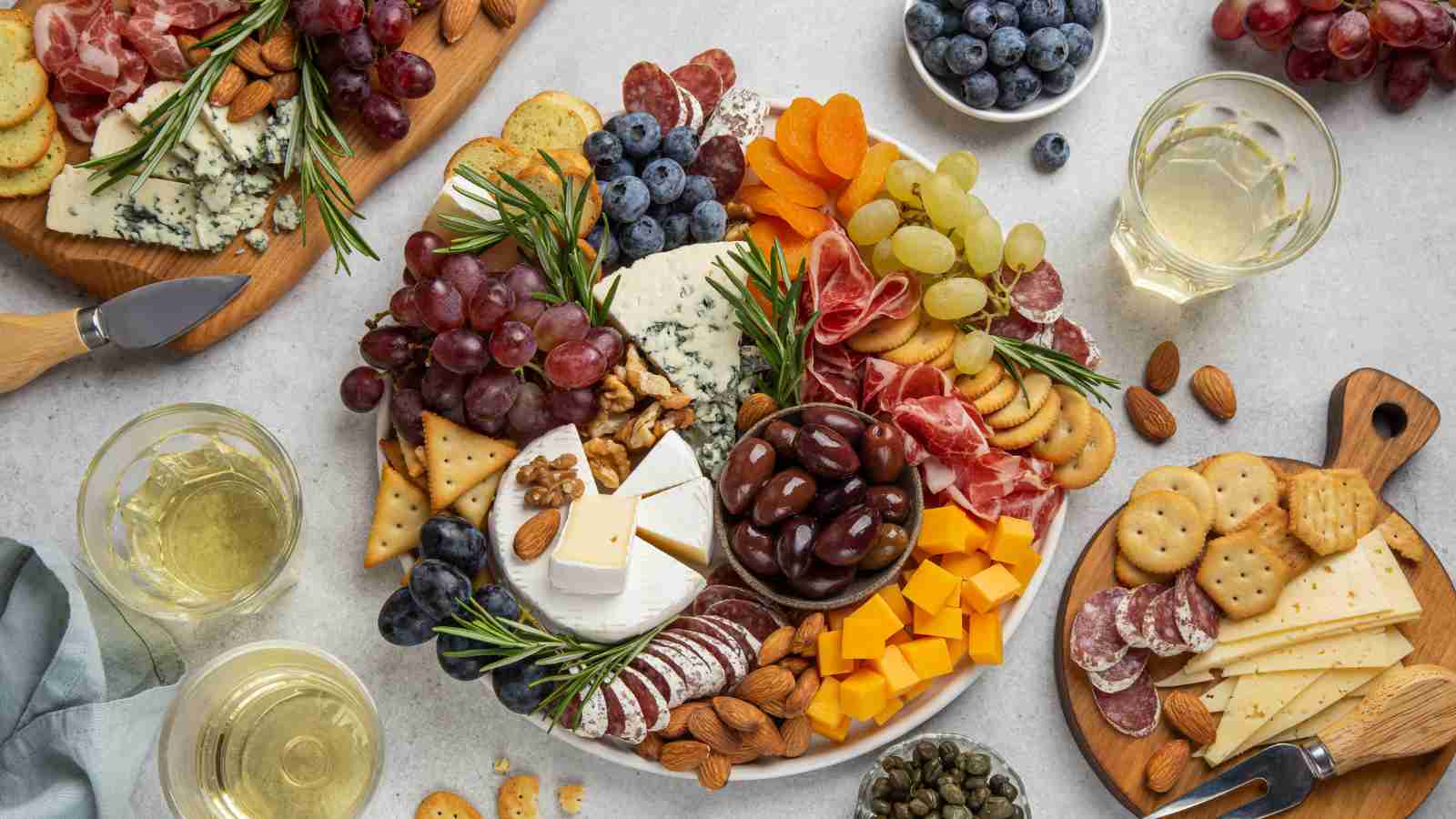An overhead view of a charcuterie board with assorted cheeses, meats, crackers, grapes, blueberries, olives, nuts, dried apricots, and glasses of white wine on a light-colored table.