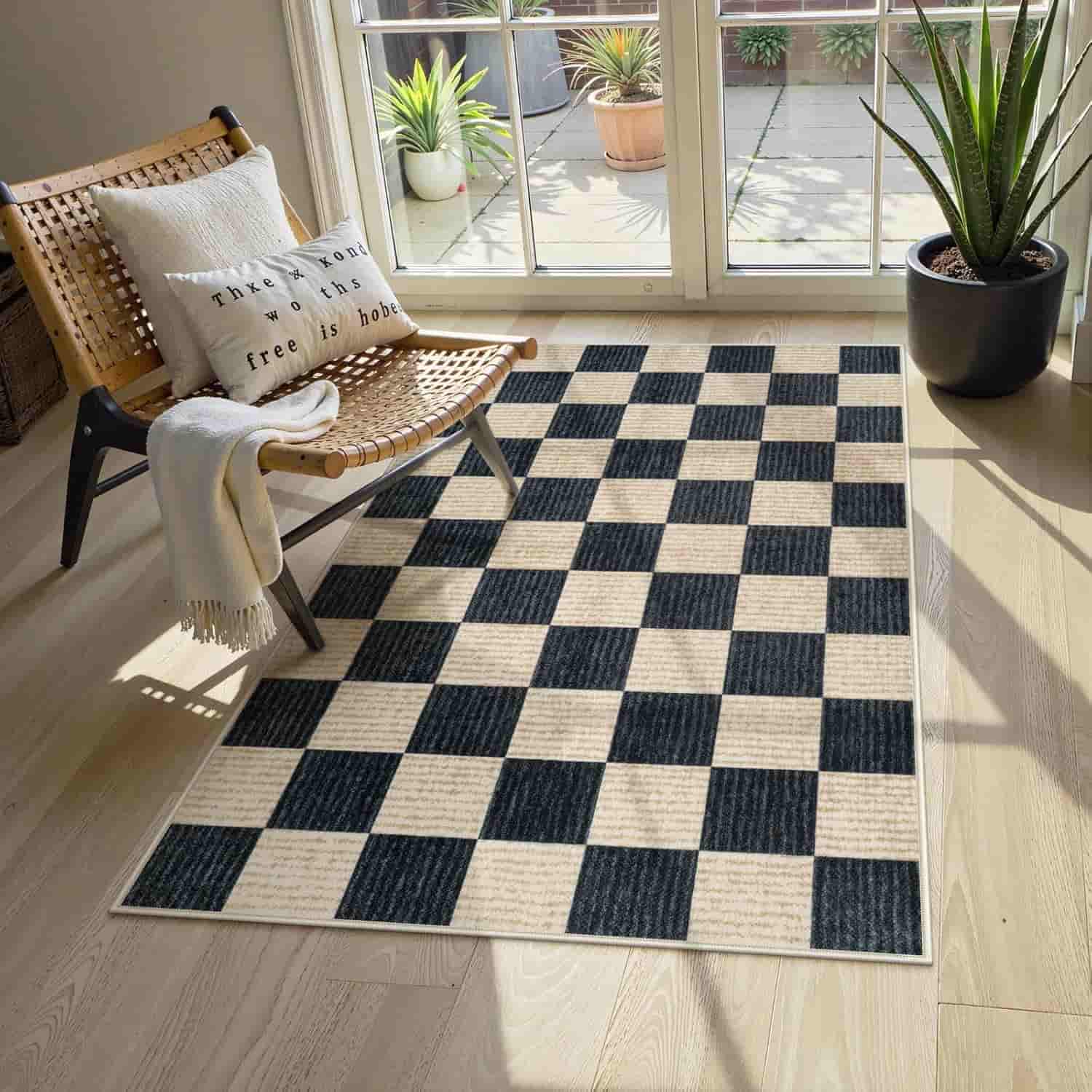 A black and white checkered rug on a wooden floor next to a woven chair with pillows and a blanket, near large windows with potted plants.