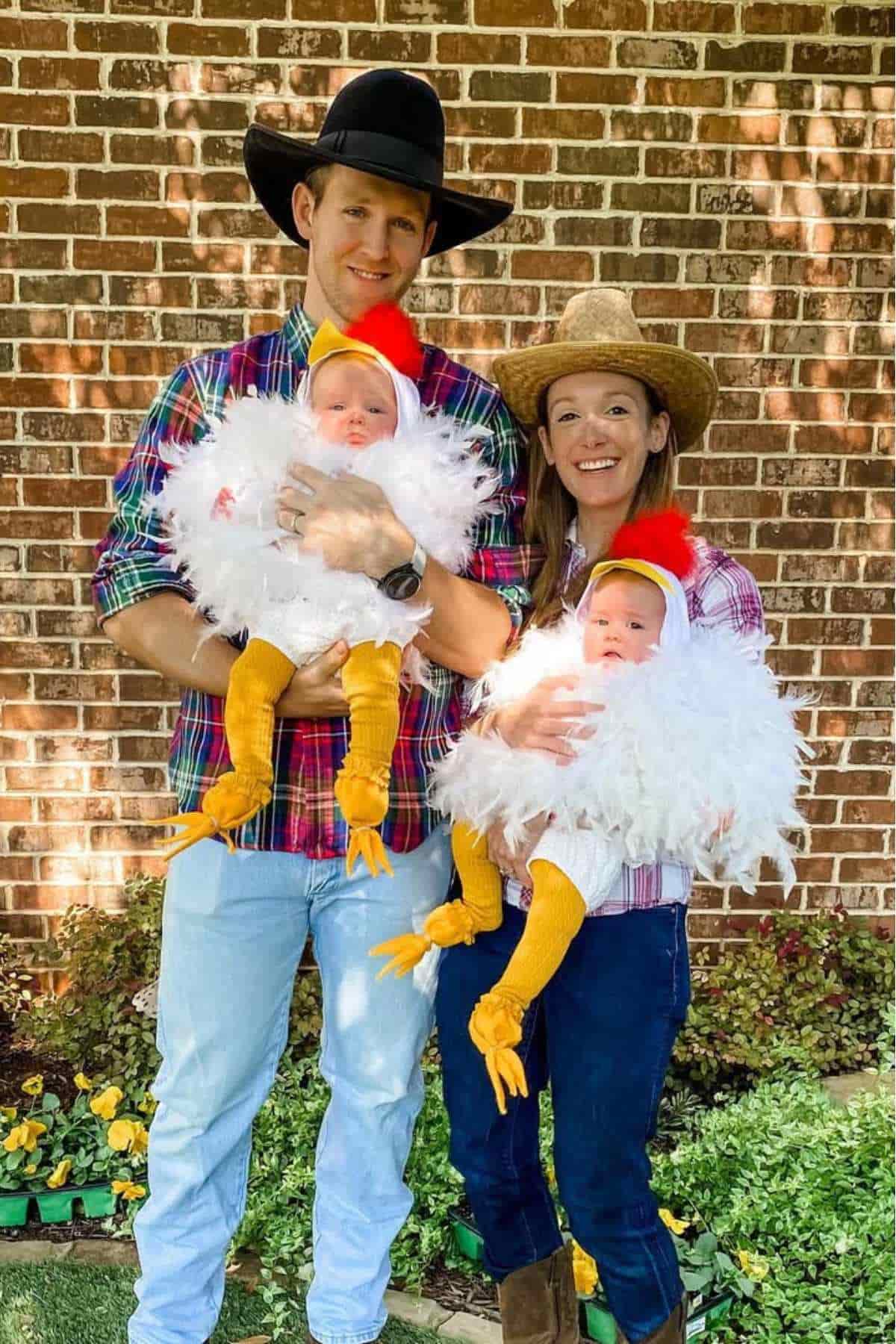 A man and woman in a farmer attire while holding two babies dressed in white feathered chicken costumes, standing outside in front of a brick wall.