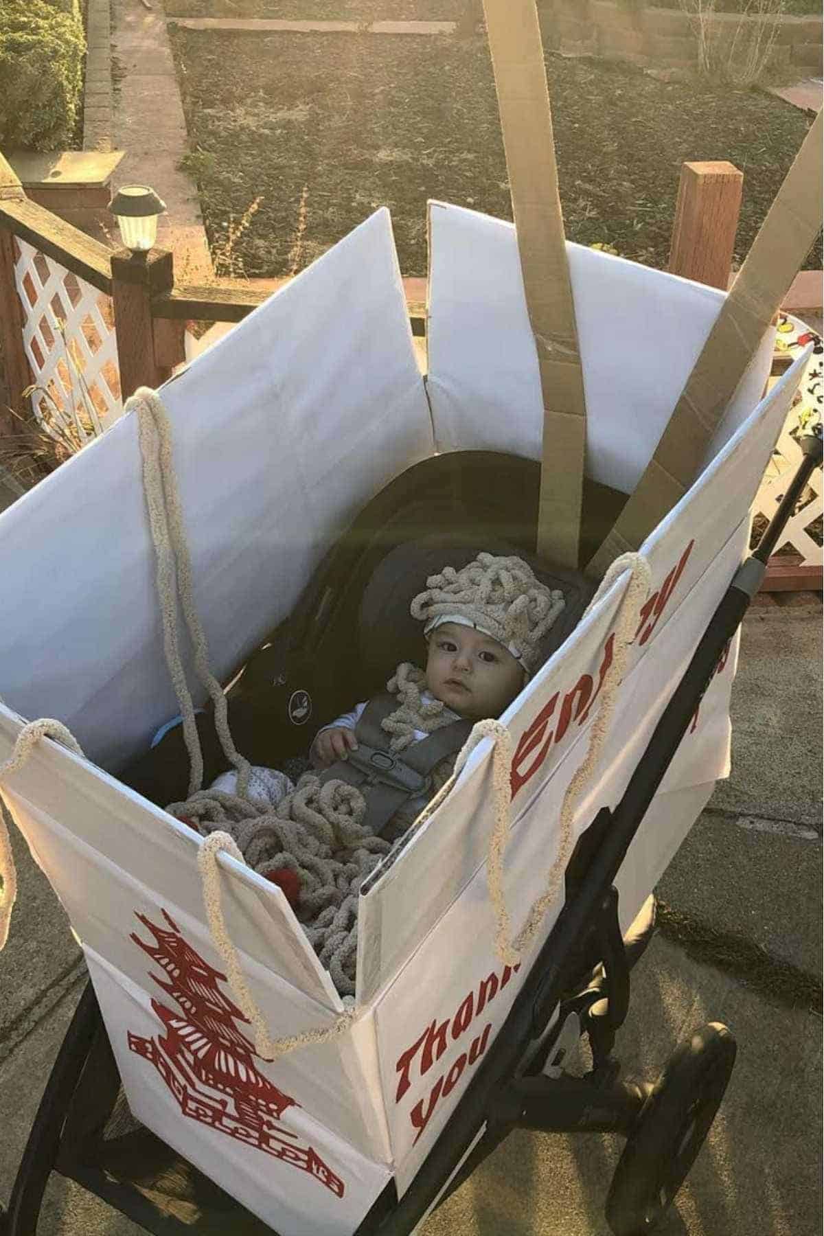 A baby in a stroller sits inside a large cart decorated as an instant noodle package, with rope resembling noodles placed around the baby.