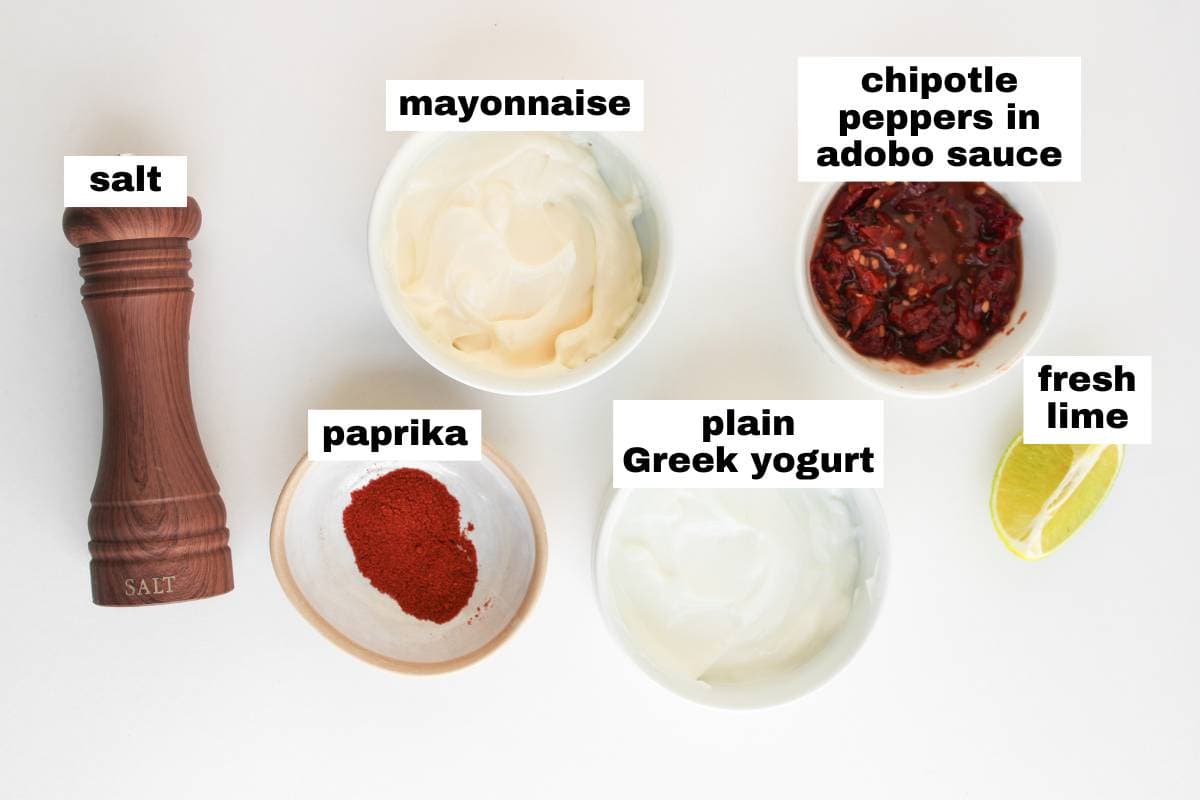 Five ingredients for a sauce are arranged on a white surface: salt, mayonnaise, chipotle peppers in adobo sauce, paprika, plain Greek yogurt, and a wedge of fresh lime.