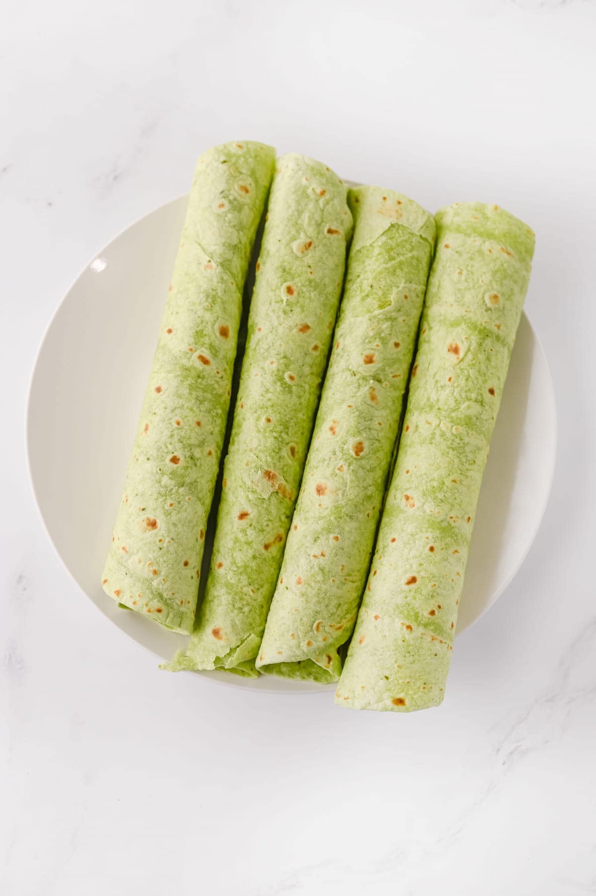 Four spinach tortillas rolled up and placed side by side on a round white plate, viewed from above on a white surface.