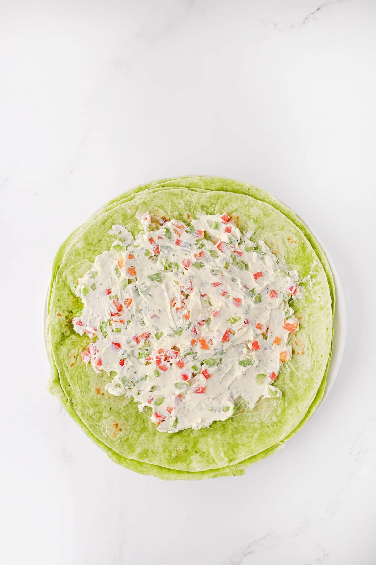 A spinach tortilla wrap on a white plate, topped with a creamy mixture containing diced red and green vegetables.