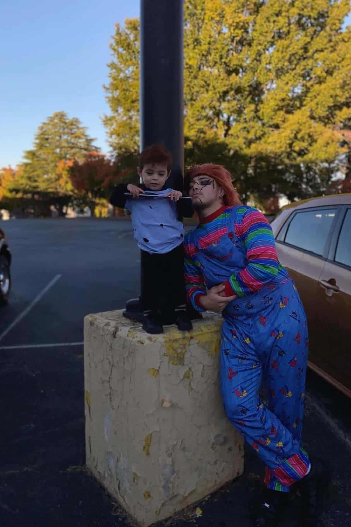 Two people in costume stand by a concrete post; one is dressed as Chucky in overalls, the other as a doll in a suit. Trees and cars are in the background.