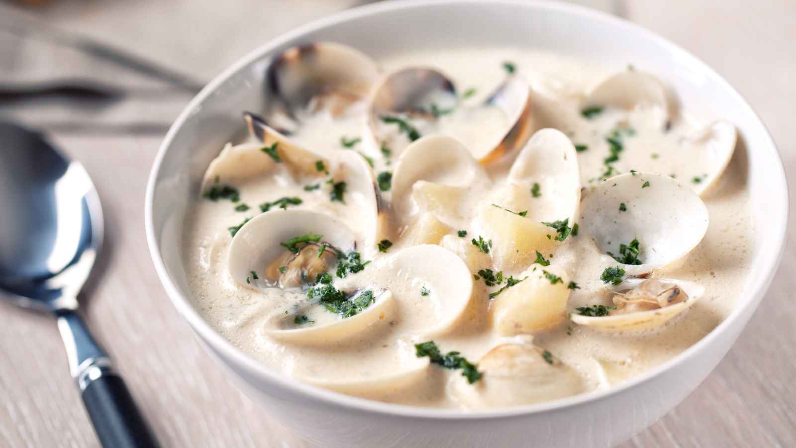 A bowl of creamy clam chowder garnished with chopped parsley, with visible clams and potatoes, next to a spoon on a light-colored table.