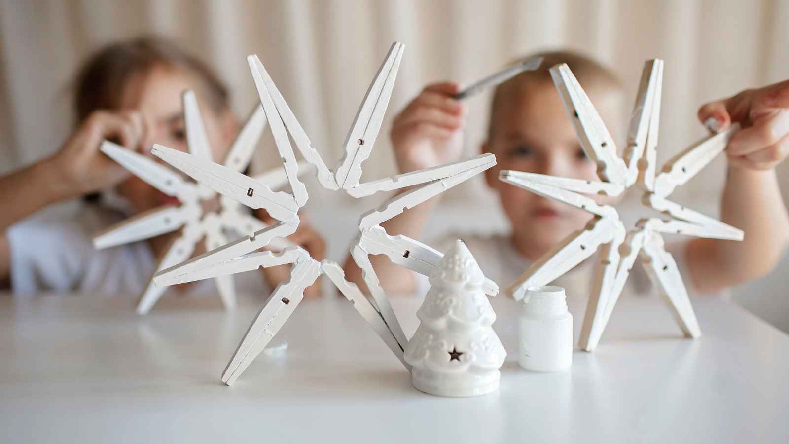 Two children hold up white star-shaped crafts made from clothespins, with a small ceramic Christmas tree and a paint jar on the table in front of them.