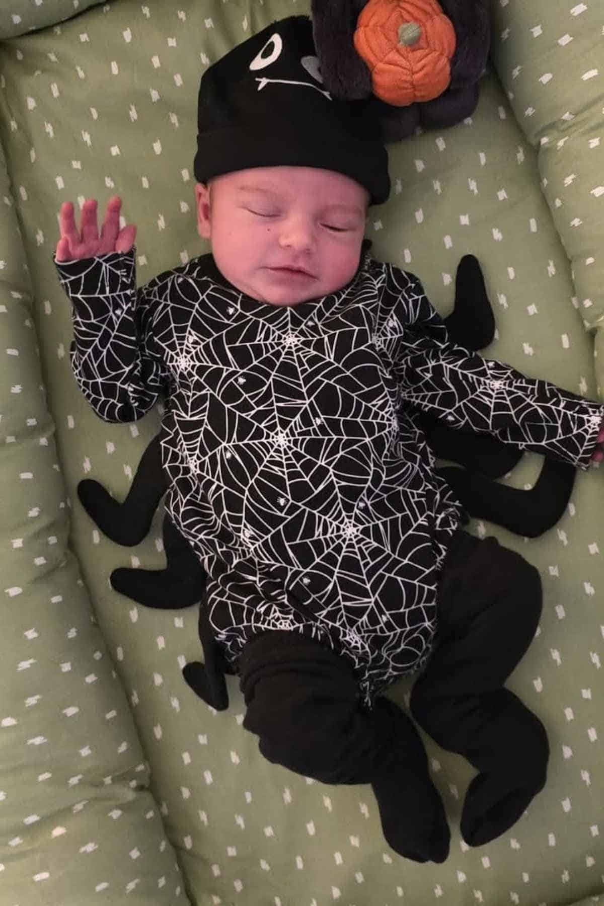 A baby dressed in a spider-themed costume with a web pattern lies on a green, dotted cushion with eyes closed.