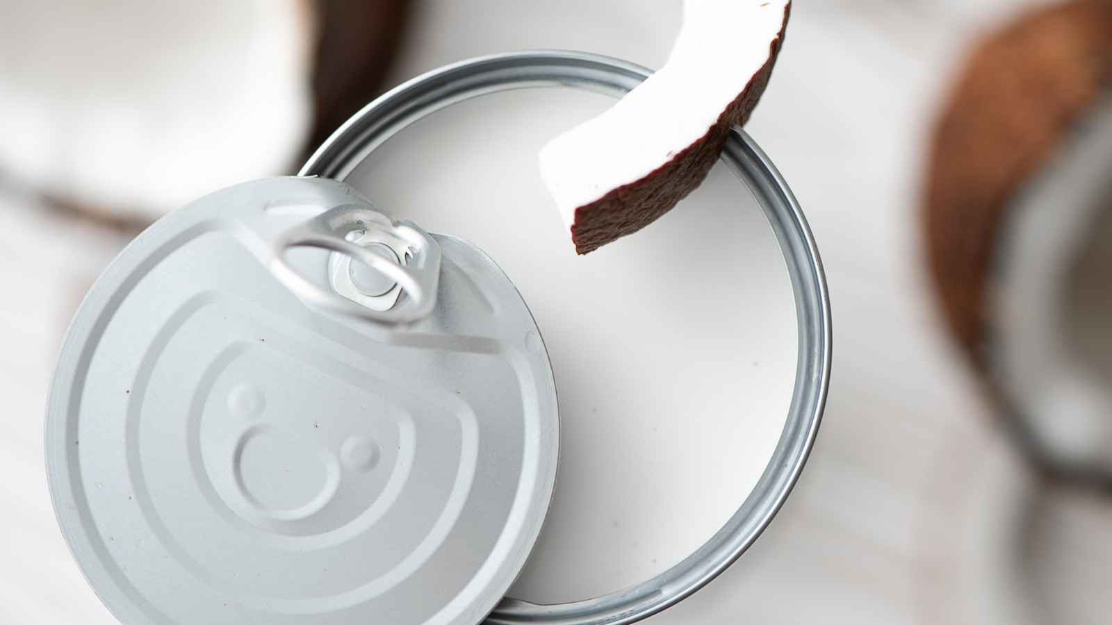 A can lid with a pull tab is open, and a piece of coconut rests on the edge of the lid. Blurred coconut pieces are in the background.