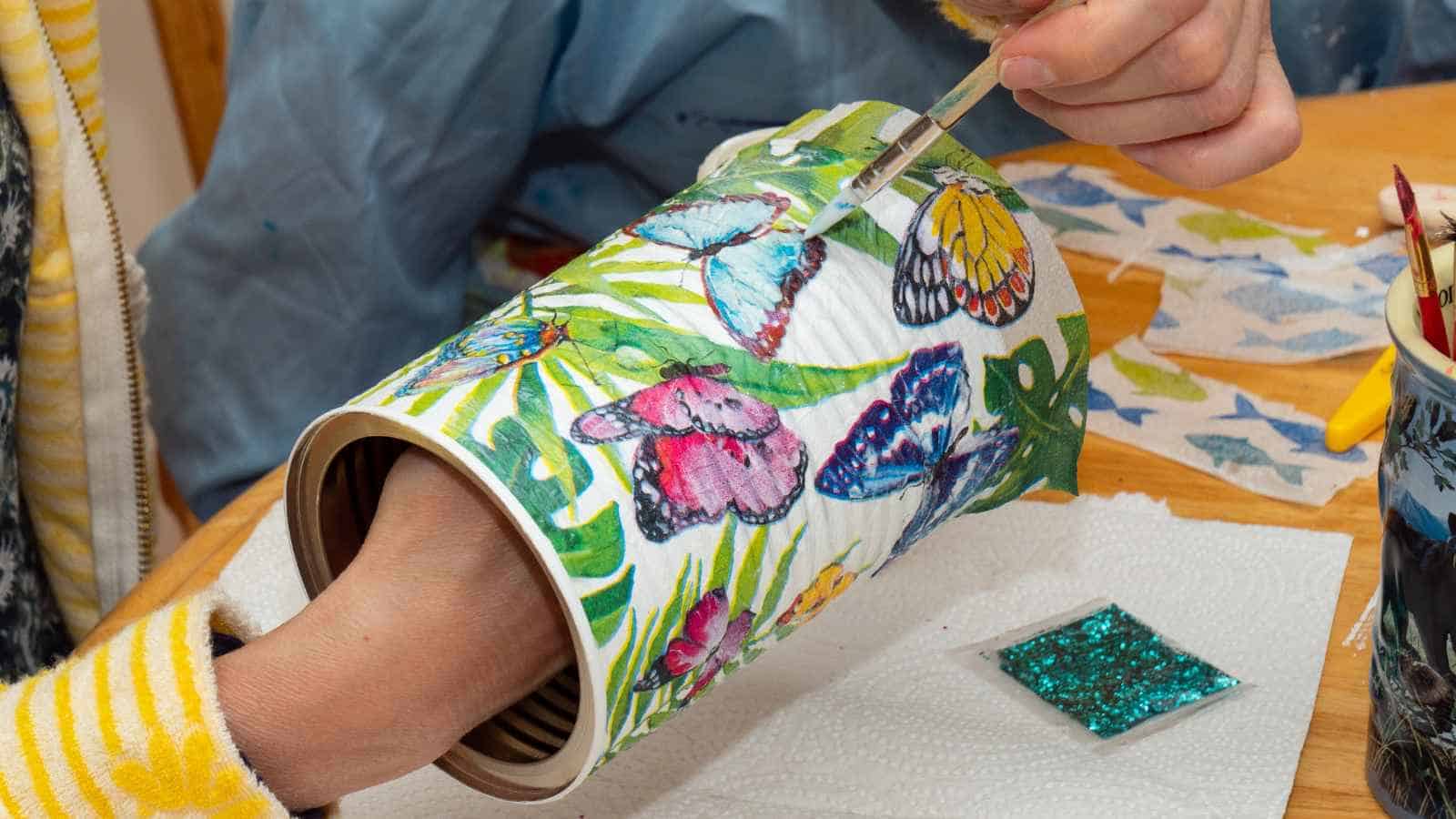 A person applies decoupage glue to a tin can decorated with colorful butterfly paper cutouts.