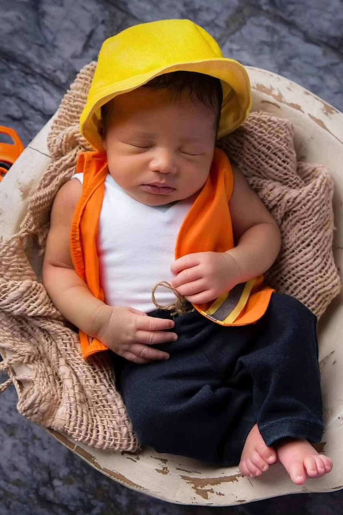 Newborn baby dressed in a yellow hat, orange vest, and white shirt, lying on a textured blanket in a bowl-shaped basket.