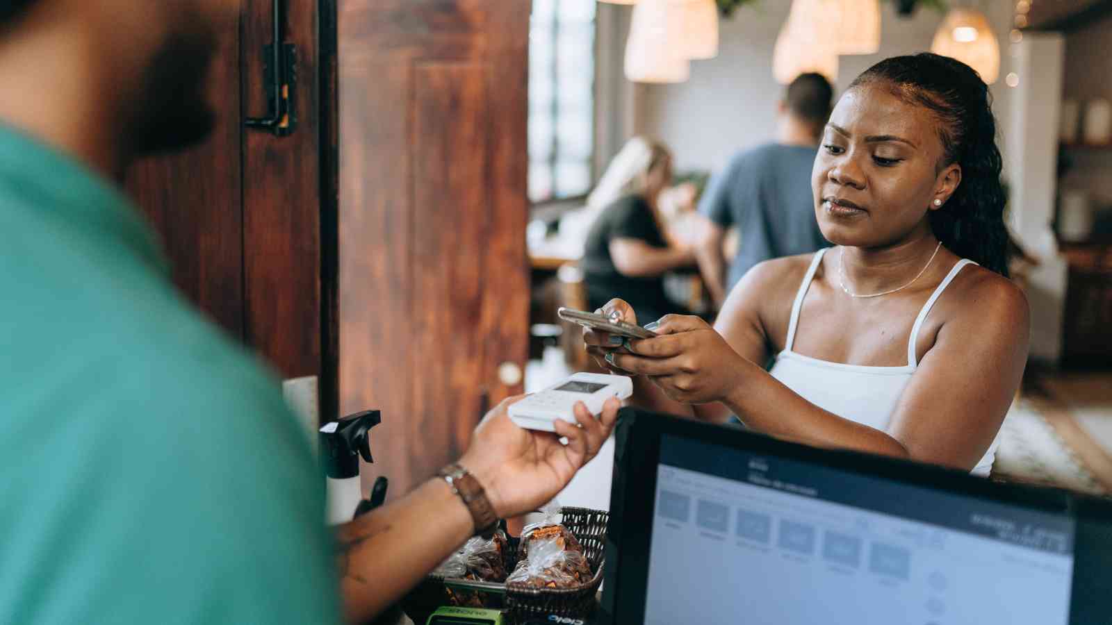 A woman holds out her phone to make a contactless payment at a counter while a person holds a card reader.