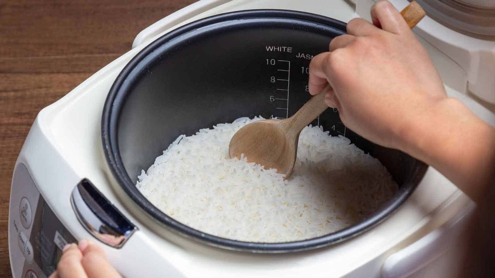 Person stirring cooked white rice with a wooden spoon inside a rice cooker.
