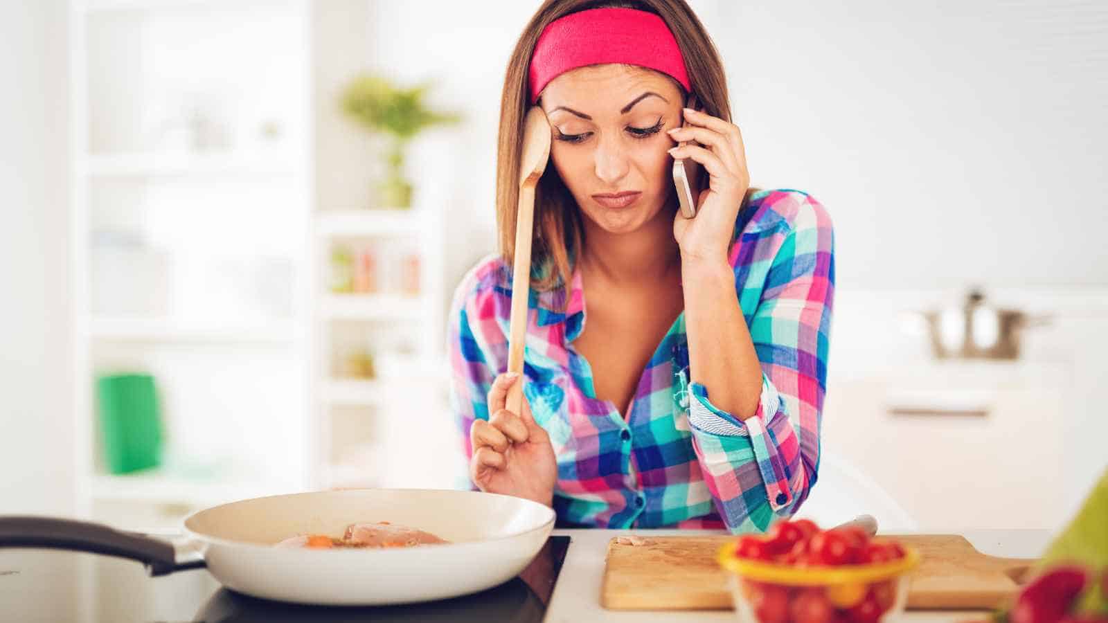 Woman in a plaid shirt talks on the phone while cooking at a stove, holding a wooden spoon, with chopped vegetables on the counter.