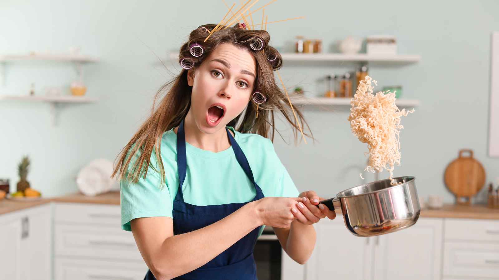 A surprised woman with hair rollers and uncooked spaghetti in her hair tosses instant noodles from a pot in a kitchen.