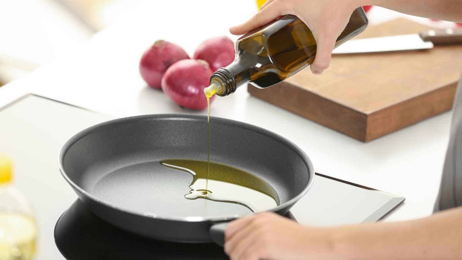 A person pours oil from a bottle into a frying pan on a stovetop, with red onions and a cutting board visible in the background.