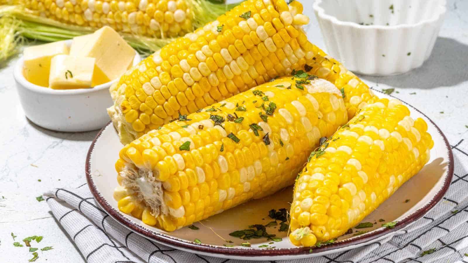 Four pieces of cooked corn on the cob garnished with chopped herbs, served on a plate beside a bowl of butter cubes.