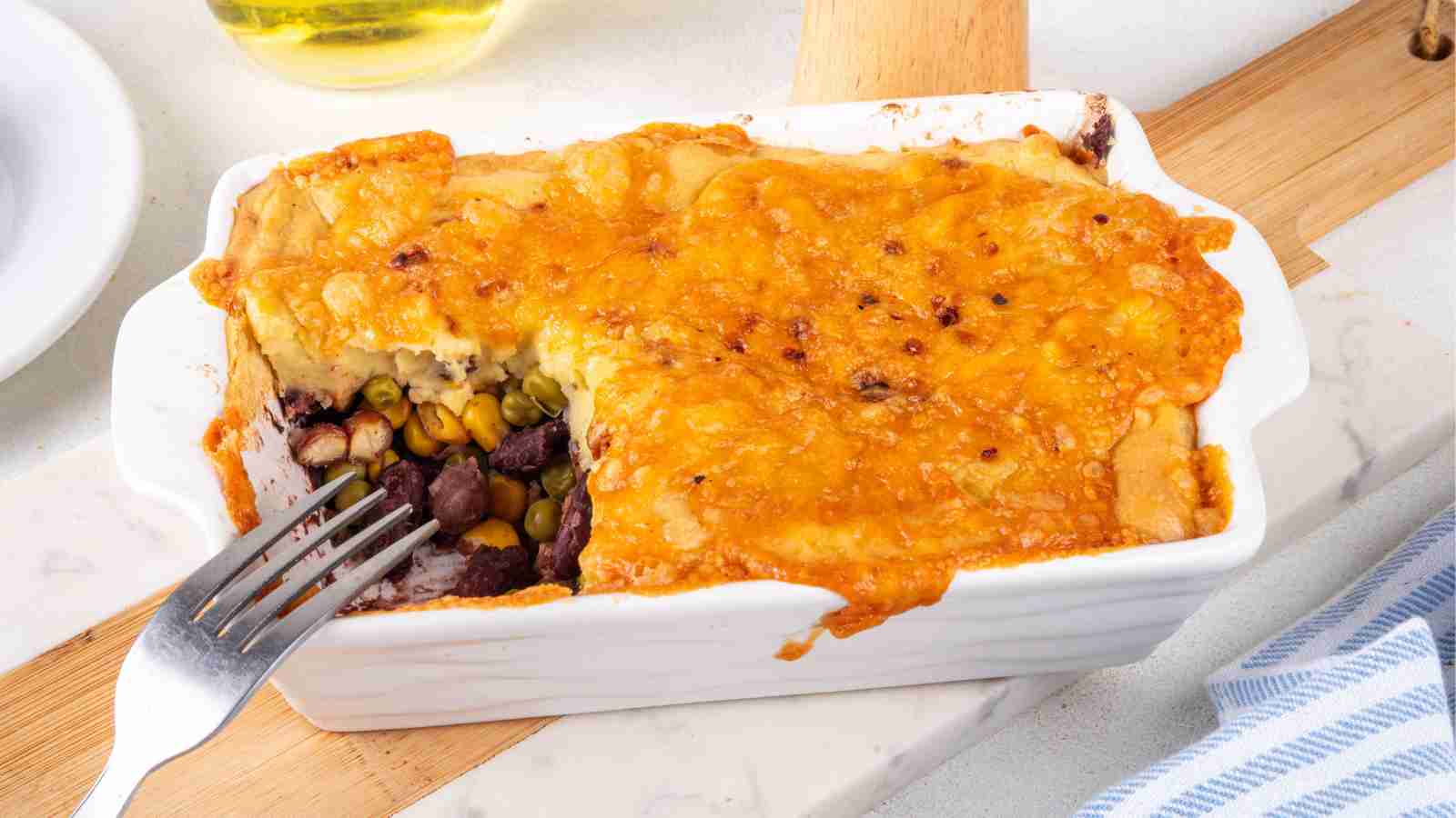 A baked casserole topped with melted cheese, with a portion removed to reveal layers of beans, corn, and vegetables inside. A fork rests on the dish.