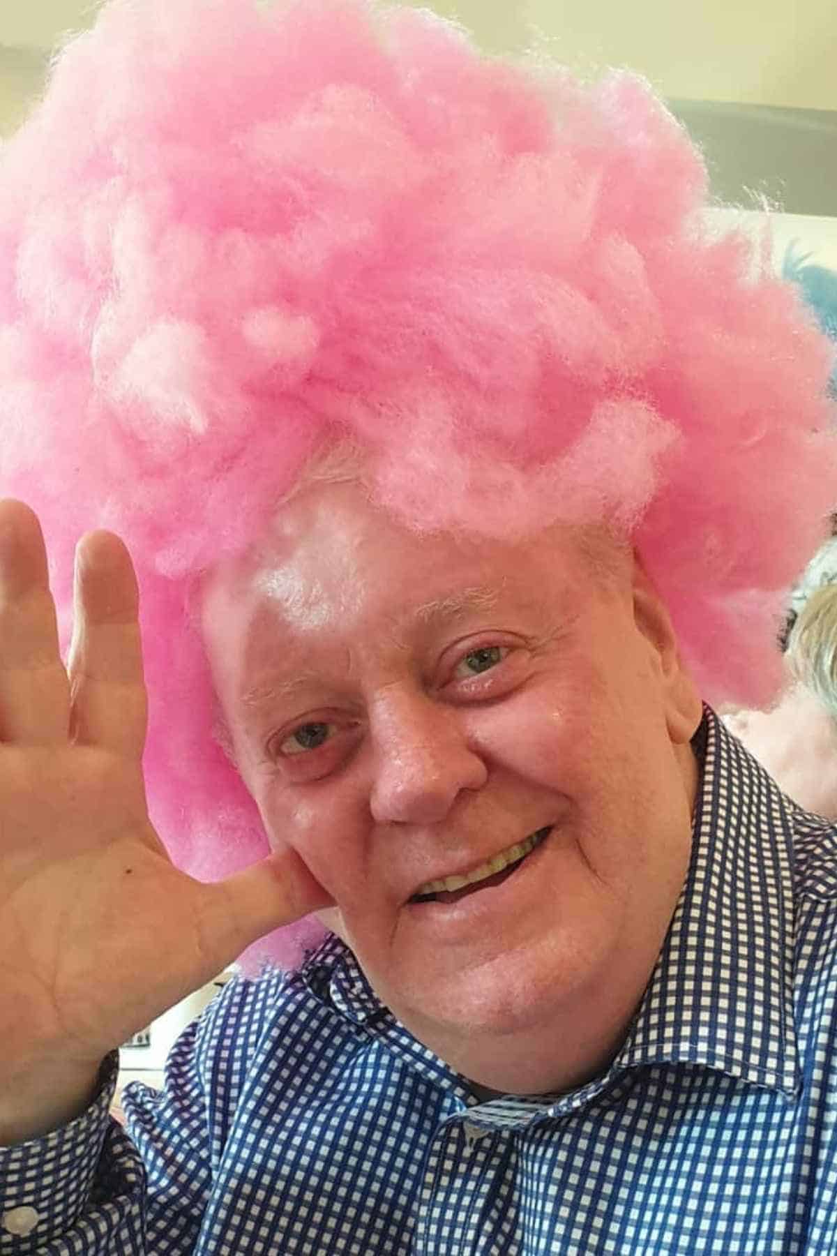 A man wearing a blue checkered shirt smiles and waves, sporting a large, fluffy cotton candy on the top of his head.