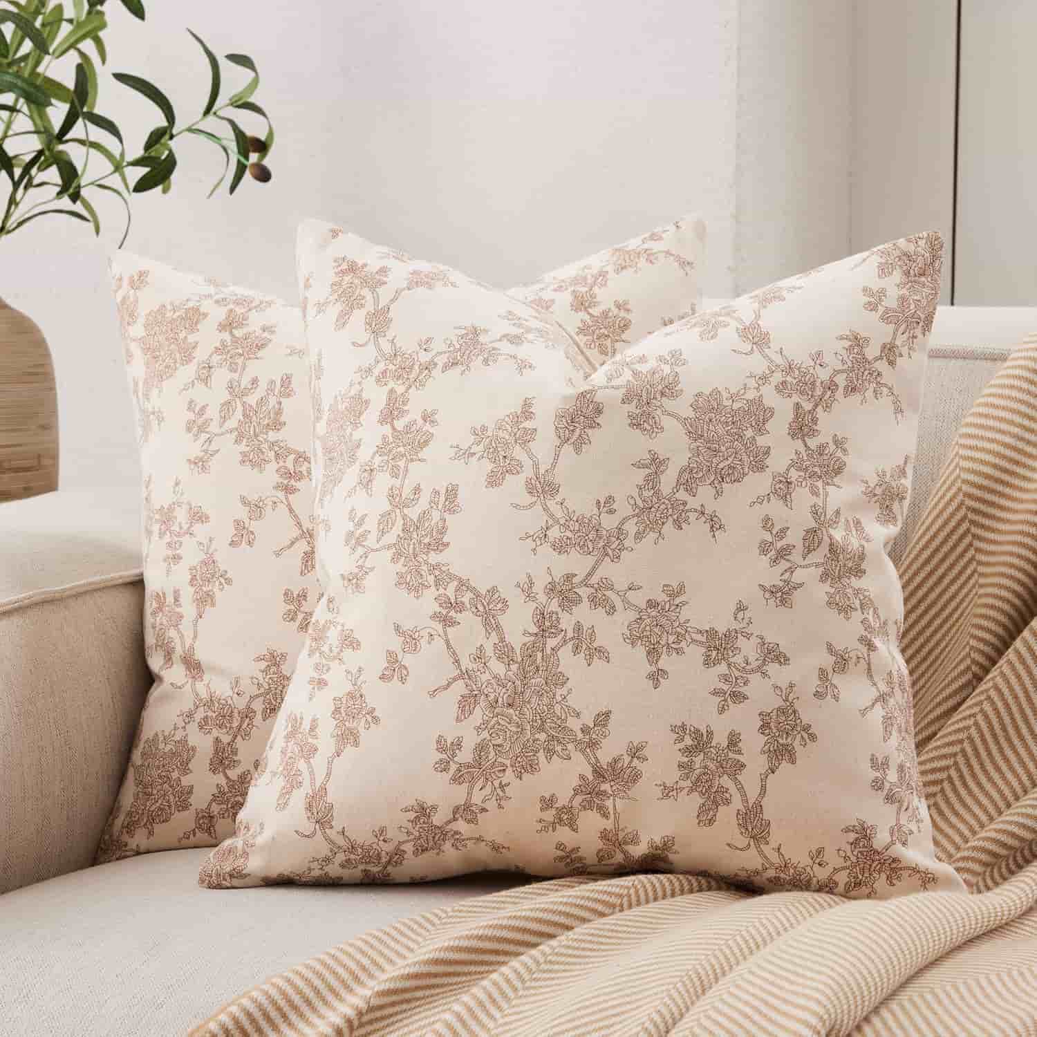 Two cream-colored throw pillows with a brown floral pattern are placed on a light beige sofa next to a beige striped blanket. A leafy plant is in the background.
