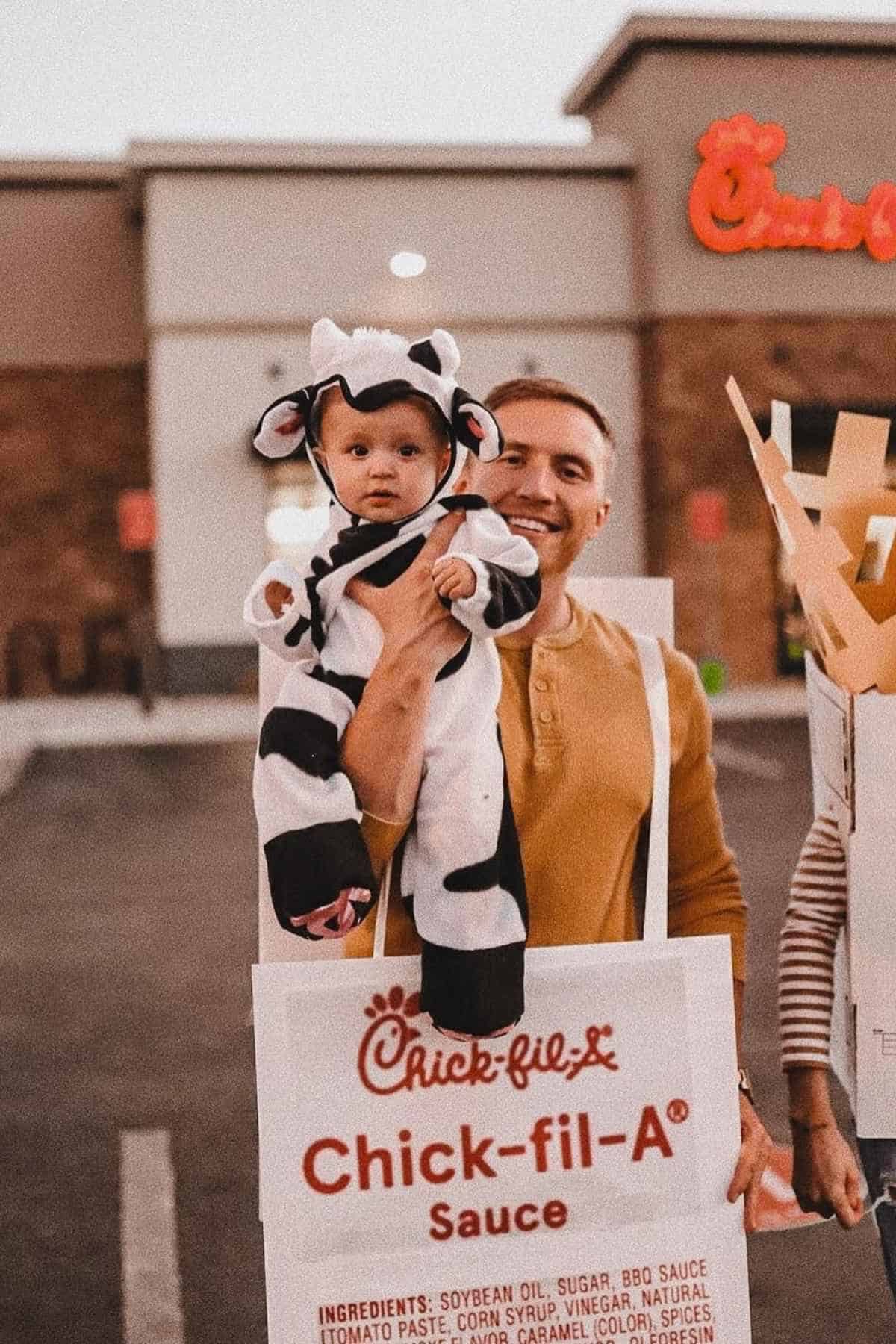 An adult holds a baby wearing a cow costume next to a large Chick-fil-A sauce packet costume, standing outside a Chick-fil-A restaurant.