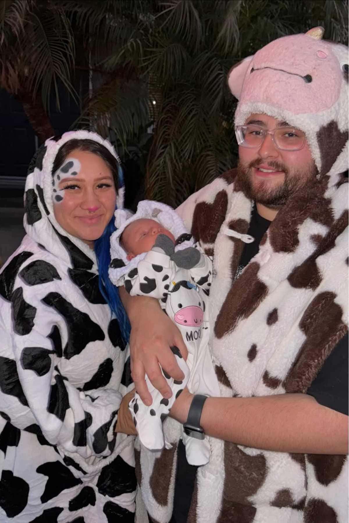 Three people, including a baby, are dressed in cow-themed costumes. The adults are standing on either side, holding the baby. There are leafy plants in the background.