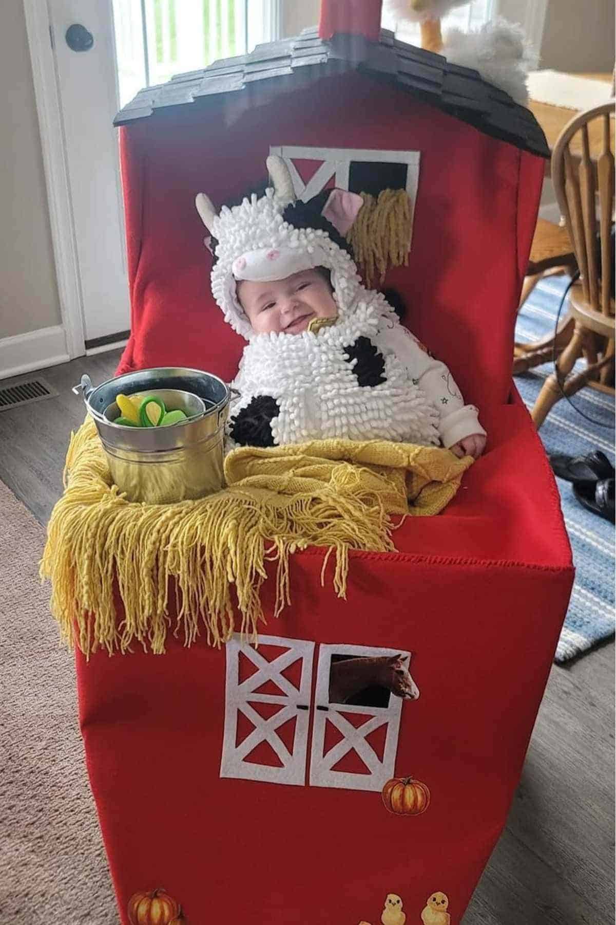A baby dressed in a cow costume sits in a red barn-themed stroller decorated with faux hay, a tin bucket, and farm animal cutouts.