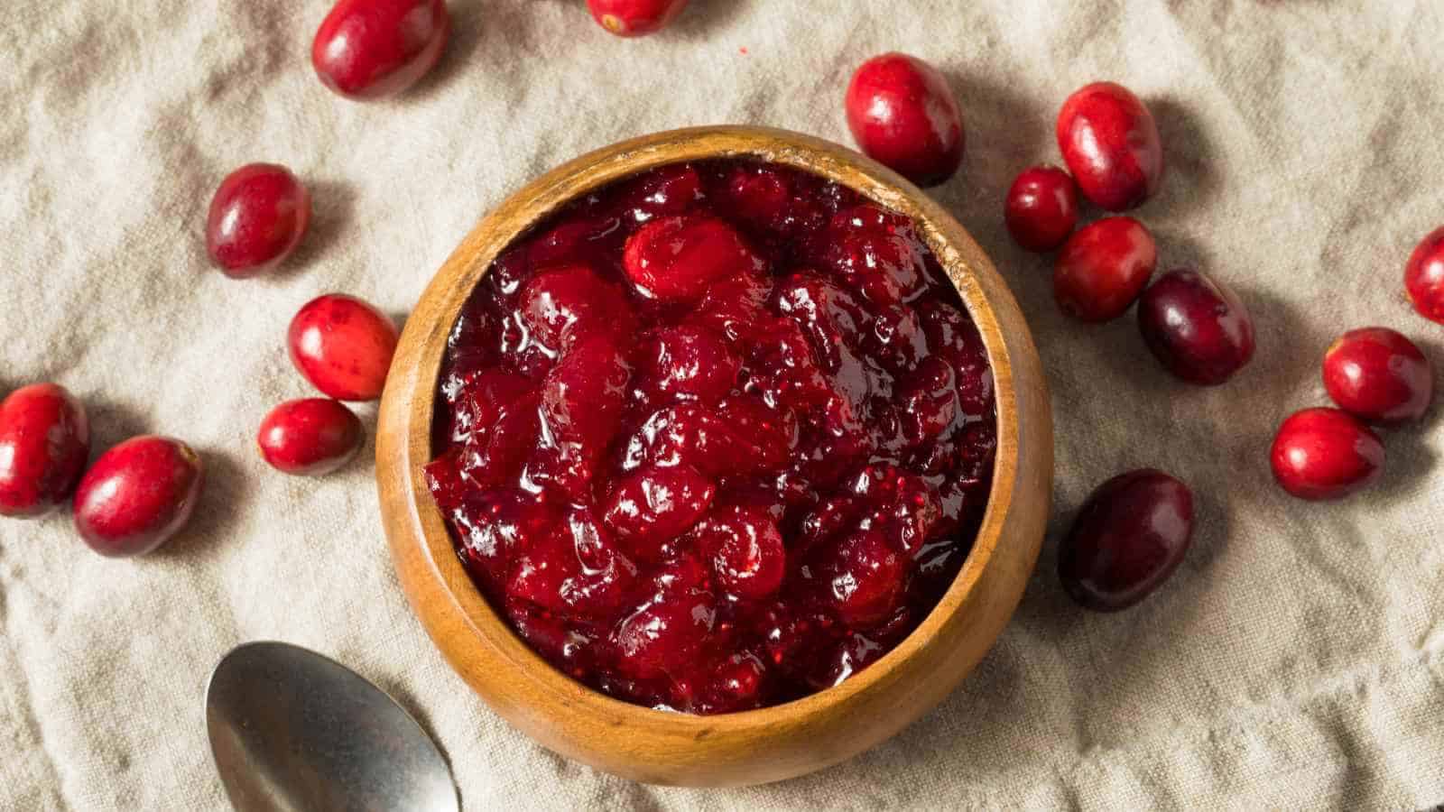 A wooden bowl filled with cranberry sauce sits on a beige cloth, surrounded by whole cranberries and a metal spoon.